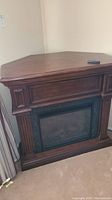 Full view of the corner electric fireplace in dark wood mantel with remote on top.