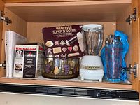Photo of shelf inside cabinet showing multiple kitchen items including Hamilton Beach blender, Wear-Ever Super Shooter box, KitchenAid hand mixer box, glass mixing bowl with fruit design, and clear measuring cup with blue clip.