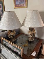 Both lamps on glass-top table with visible cords and picture frames in background.