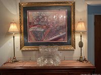 Pair of matching lamps placed on striped fabric covering a wooden table, with a painting in a gold frame behind them.