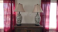 Pair of matching table lamps with ceramic bases decorated with colorful bird and floral patterns on a wooden chest, beige lampshades, positioned in front of windows with red curtains.