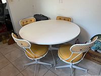 Front view of the round white Formica table with four swivel chairs around it, showing mustard-yellow padded seats and cane backrests.