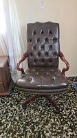 Front view of vintage brown leather office chair with tufted backrest, wooden curved armrests, and nailhead trim along seat edge.