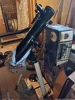 Full view of Meade Polaris 114EQ-D telescope mounted on aluminum tripod with black mount head, standing next to original box in basement setting.