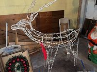 Full side view of the white wire frame reindeer decoration with string lights tied around the frame and red fabric around the neck, positioned indoors on concrete floor.