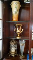 Three vases and a candle stand on wooden shelves; includes yellow ceramic vase with white daisies, gold-tone vase with handles, candle stand with crystal beads, and tall fluted vase with gold decoration.