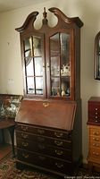 Front view of vintage wooden secretary desk and matching hutch with glass paneled doors, brass handles, and carved crown top.