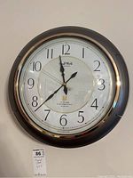 Full front view of the round wall clock with black frame, gold trim, white face with black numbers and hands.