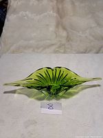 Green Murano glass centerpiece with an abstract organic shape, ribbed texture, and elongated tips, placed on a white textured surface with a number 8 tag in front.