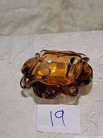 Front angle view of amber Murano art glass ashtray with ruffled edges and smooth finish on white fabric background with lot number 19 tag.