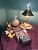 Overview of entire lot on burgundy tablecloth showing brass lamp, ceramic vase, clock, wooden box, porcelain doll, framed wood carvings, and small figurines