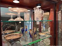 Wide view showing all items in the lot including the clear glass cake stand, cut glass pitcher, silverplate bowl, cobalt blue vases and small bowl arranged on glass shelves.