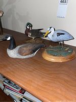 Image showing three hand carved waterfowl wood figurines displayed on a wood surface: a large duck, a blue heron on an oval wooden base with natural elements, and a smaller duck on a stump.