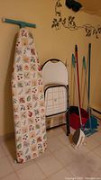 Ironing board, folding chair, mops, broom, dustpan arranged against wall on tiled floor
