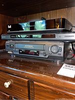 Stacked Sony electronic devices including a DVD/CD player and VCR on a wooden surface with a Bell receiver also present