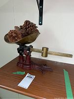Full view of vintage Fairbanks weigh scale with red cast iron base, brass bowl holding pine cones, and brass sliding weight on measuring arm.