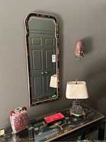 Mirror hanging on a wall next to a wall lamp and red and white vase placed on a decorated black lacquered surface beneath it.