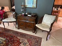 Pair of two caned wood-framed chairs with beige cushions positioned on either side of a wooden cabinet, showing carved back detail and cane weaving.