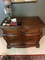 Front view: Shows wooden sideboard cabinet with curved details, drawer with brass handles, and two cabinet doors with matching brass handles. Top surface holds miscellaneous small items and a vintage lamp.