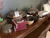 Wide shot of lot showing two hexagonal silverplate covered serving dishes with glass inserts, coaster sets in boxes, pottery bowl, trinket boxes, napkin rings, matches, and watch on wooden furniture surface.