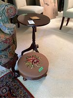 Full view of oval side table with tripod legs next to footstool with floral needlepoint upholstery.