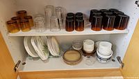 Glass shelf with amber to brown gradient glasses on top shelf and clear, white, and patterned plates and small bowls on bottom shelf.