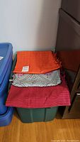 Three fabric remnants stacked on a plastic tote: orange checked, light blue with diamond pattern, deep pink striped.