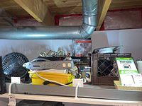 Lot overview showing table top fan, power bars, coiled cords, LED security light box, smoke alarms, and work light box on yellow bin under basement ceiling pipes.