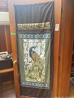 Full view of brown fabric wall hanging with colorful peacock and branch print, with decorative gold and feather motifs at top.