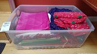 Plastic bin holding multiple folded silk saris and shawls, showing pink, blue, and red fabrics through translucent container.