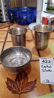 Photo showing two silver cups and one stamped silver bowl on a patterned table surface.