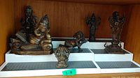 Five brass Hindu statuettes arranged on white plastic display stand on wooden shelf, showing various postures and sizes labeled 3-8 inches.