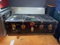 Full view of the rectangular black vintage steamer trunk showing brass locks and metal rivets on corners and edges.