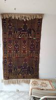 Full view of large rug hanging on wall with geometric and animal motifs and fringe edges, rest of room partially visible.