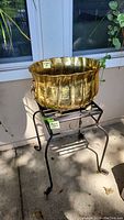 Full view of brass planter placed on black metal plant stand outdoors, showing overall shape and condition.
