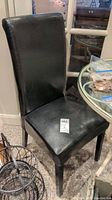 Black faux leather dining chair angled showing front seat cushion and high backrest with tag on seat
