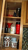 Wooden kitchen cabinet shelf with red funnel, black French coffee press, red and black travel mug, white floral mug, brown and white ceramic mugs, and dark bottles.