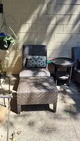 Outdoor dark brown faux wicker chair with matching ottoman and patterned pillow shown on patio floor against brick wall. Natural lighting, shadows from nearby plants.