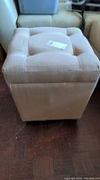 Front and top angled view of beige square tufted fabric ottoman with button accents and wood legs on a dark wood floor.