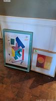 Two of the framed wall art pieces leaning against a wall on a hardwood floor: one larger with geometric shapes in green frame, one smaller with circular design in gold frame
