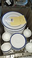 Stack of white ceramic dinner plates with double blue rim lines and matching cups and saucers arranged inside a crate.