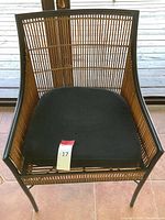 Front view of a chair with a black metal frame, bamboo woven sides and back, and a black upholstered seat cushion placed on a tiled floor.