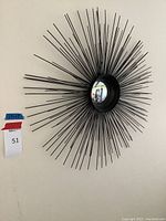 Full view of the decorative metal sunburst funhouse mirror mounted on a wall showing the radiating metal rods and convex mirror at center.
