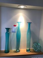 Photo of three tall blue glass decorative bottles and one small clear blue glass bowl on a wooden shelf under spotlight lighting.