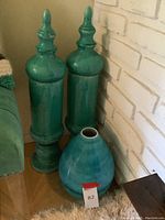 Two tall turquoise blue ceramic vases with finial tops, standing side by side next to a white brick wall with a shorter round vase in front of them on the floor.