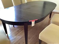 View of the oval dark wood dining table with part of the removable leaf extended, showing straight legs and smooth finish.