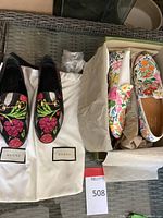 Overhead view of both pairs: black embroidered loafers on dust bags with Gucci tags; ivory floral print loafers in box with tissue paper.