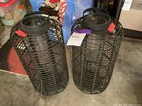 Two black metal outdoor lanterns standing side-by-side on a concrete floor with sale tags. Each lantern has a cage-style metal frame and a handle on top.