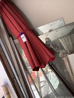 Photo showing the closed reddish orange outdoor umbrella leaning against a wall indoors with a visible metal pole and crank mechanism.