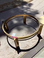 Circular metal table frame with brass/gold finish shown outdoors on concrete, pine needles on surface